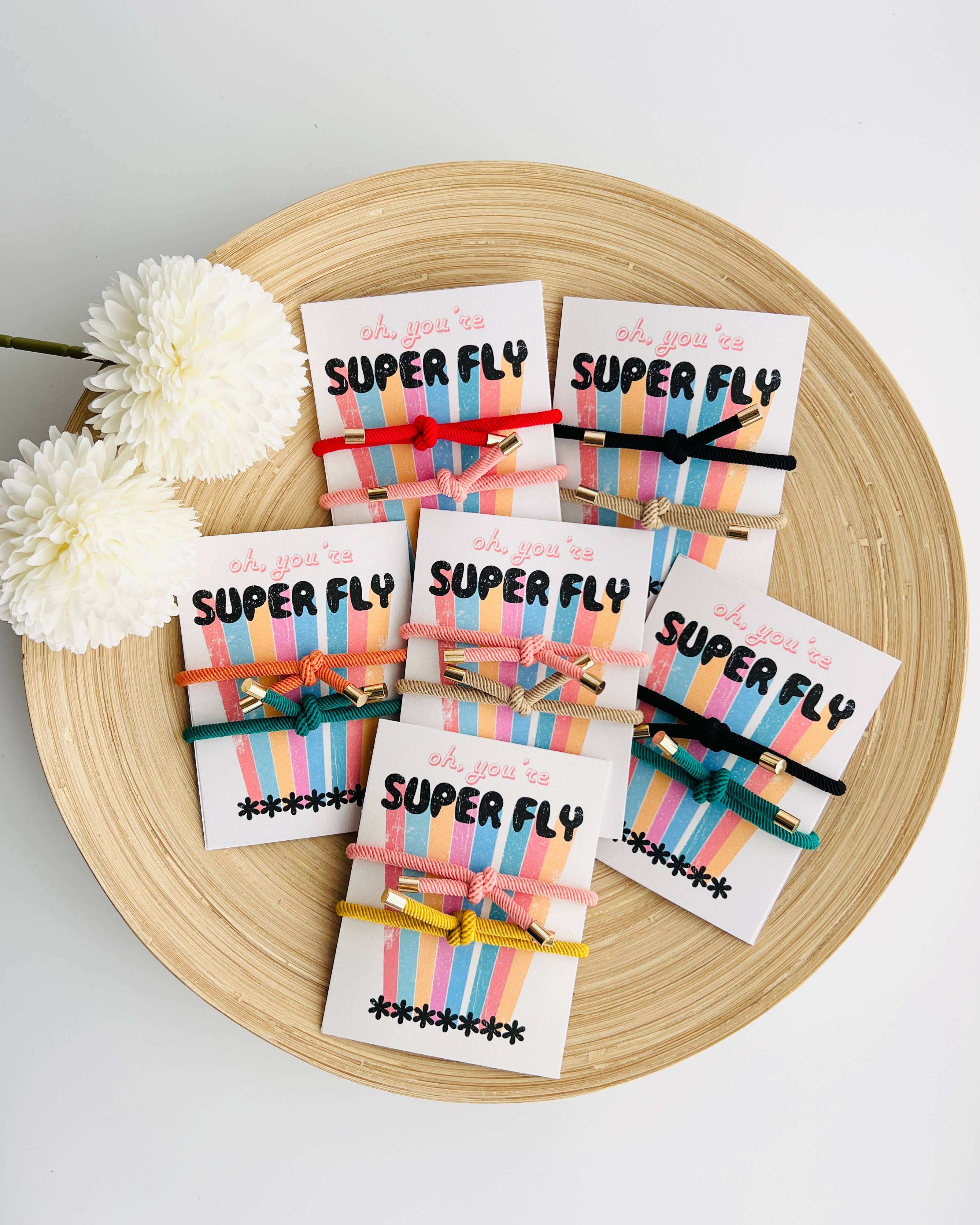 Six small gift cards wrapped in colorful striped paper and tied with wooden clothespins, arranged on a round wooden tray with white flowers. The cards read 'oh you're SUPER FLY' with star patterns.