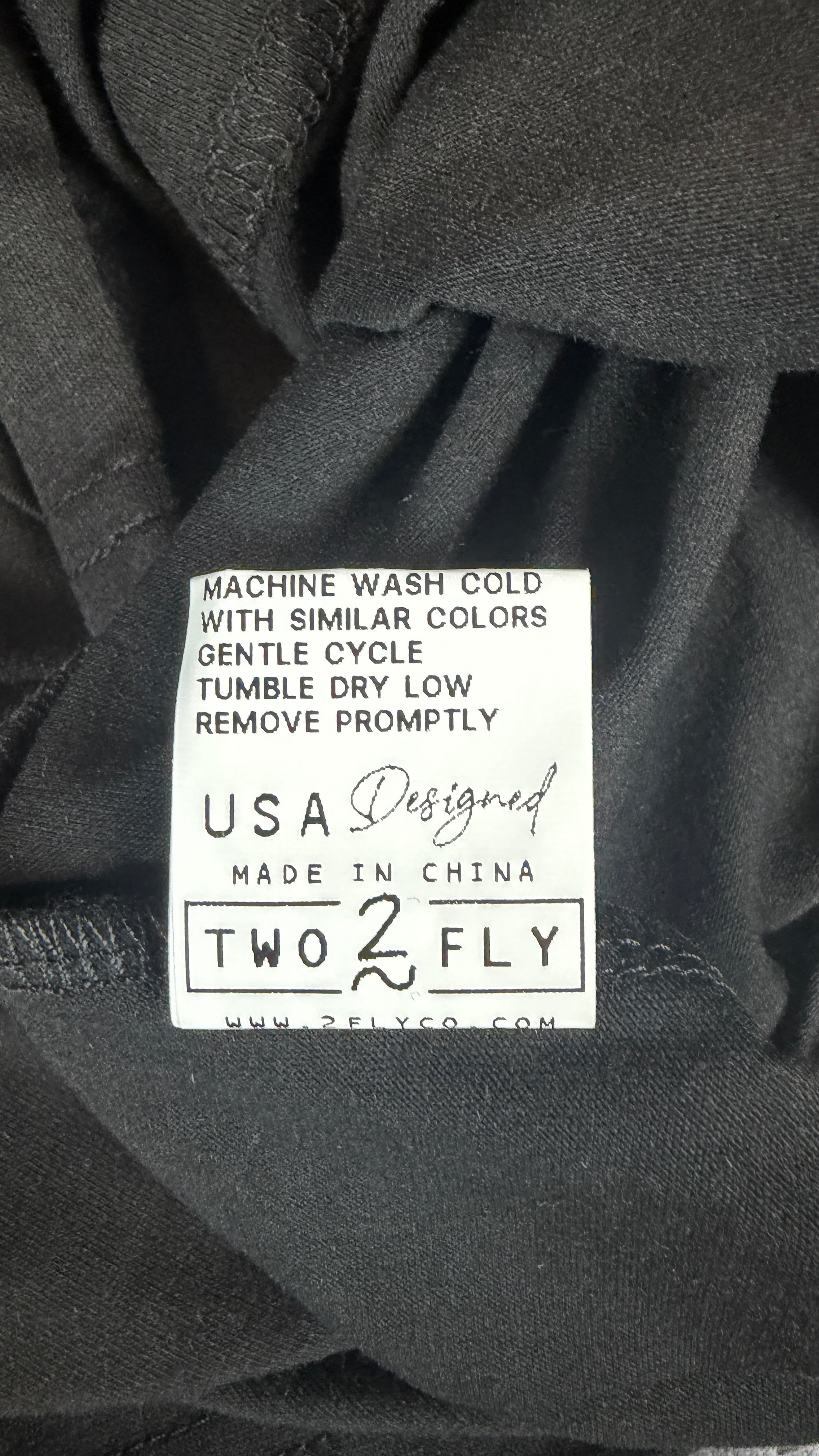 Close-up of a black garment label reading 'MACHINE WASH COLD WITH SIMILAR COLORS GENTLE CYCLE TUMBLE DRY LOW REMOVE PROMPTLY USA Designed MADE IN CHINA TWO TFLY' on dark fabric.