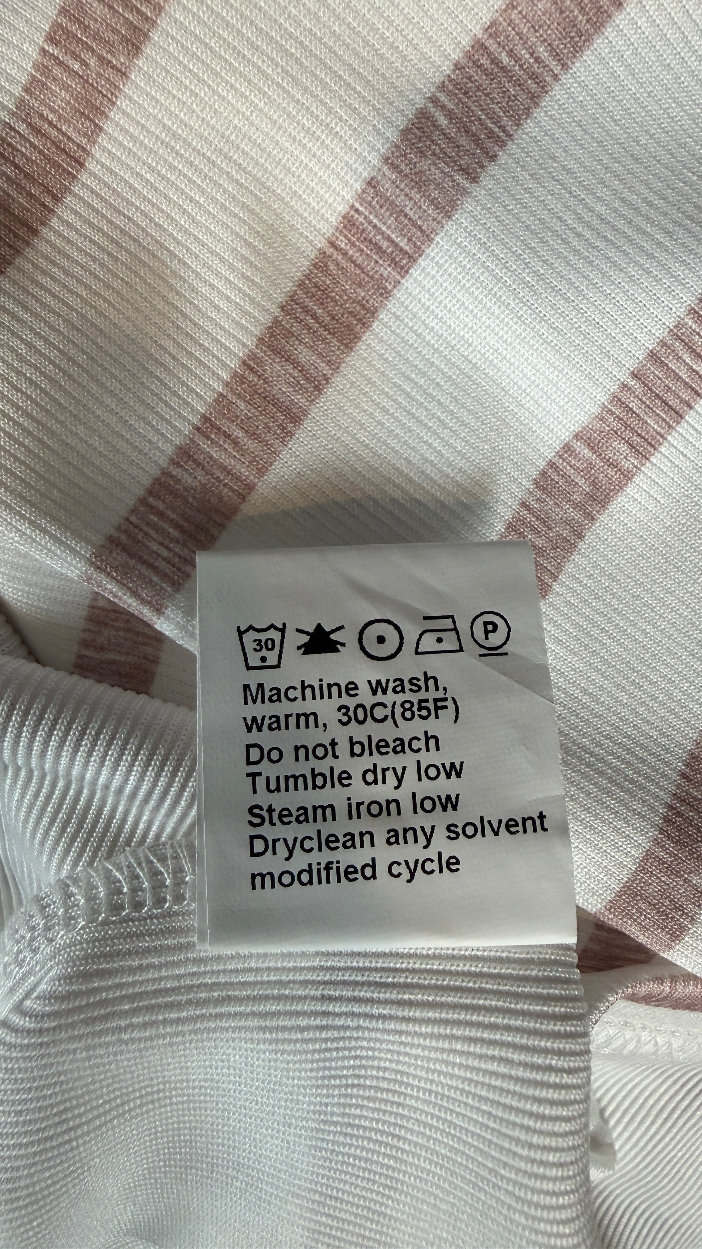 A close-up of a white garment tag with care instructions, placed on a ribbed white fabric with dusty pink stripes.