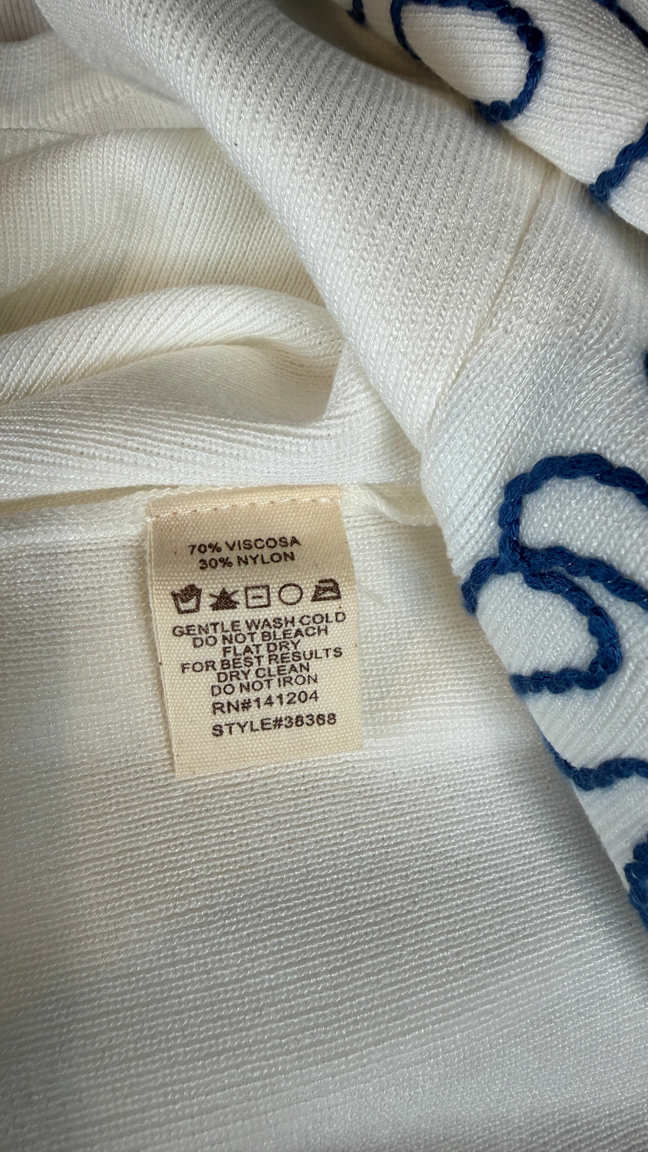 Close-up of a cream-colored knit garment with a visible sewn-in care label, showing blue embroidered or printed wave-like patterns along the edge.