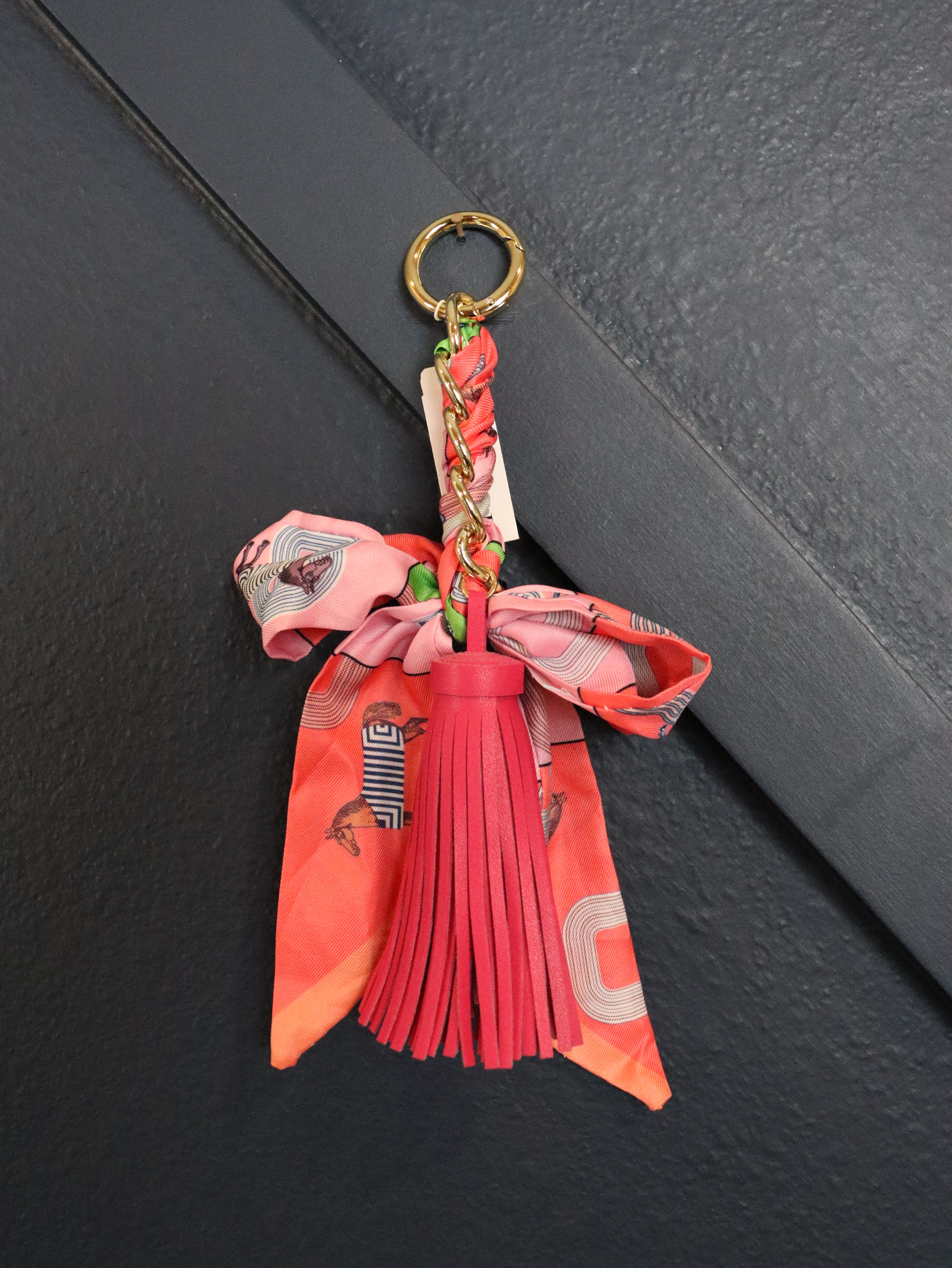 A decorative keychain featuring a coral pink and orange patterned silk scarf bow with a pink tassel, attached to a gold metal ring 